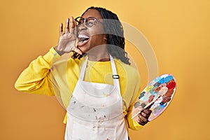 African woman holding painter palette shouting and screaming loud to side with hand on mouth