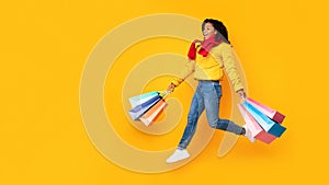 African Woman Holding Shopping Bags Running In Mid-Air, Yellow Background