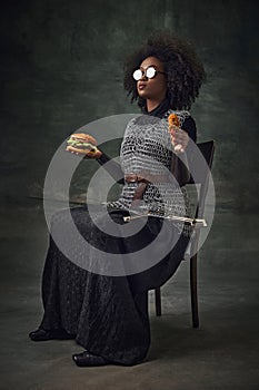 African woman, medieval warrior in chainmail, sunglasses, holding burger and chicken nuggets against dark vintage