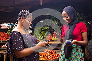 African woman shopping food stuff in a local market paying by doing mobile transfer via phone for a trader