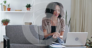 African woman sit on couch wear headset use laptop having distant communication, telework provide support to client by