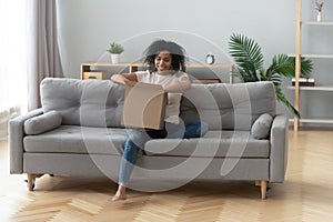 African woman sitting on couch unpacking received parcel feels happy