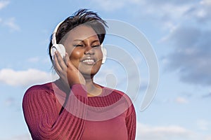 African woman sitting listening to music through headphones, with the blue sky in the background.