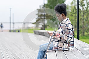 African woman skater reading sms message in smartphone app sit with longboard on bench in city park