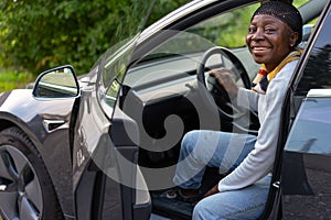 African woman smiles at camera and is sitting in electric car with open door.