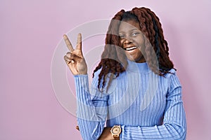 African woman standing over pink background smiling with happy face winking at the camera doing victory sign with fingers