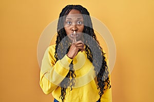 African woman standing over yellow background asking to be quiet with finger on lips