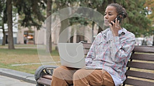 African Woman Talking on Phone and using Laptop while Sitting Outdoor on Bench