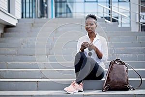 African woman using smart phone while sitting on white stairs outdoors.