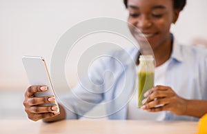 African Woman Using Smartphone Drinking Smoothie In Kitchen At Home