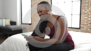 African woman using smartphone sitting on bed looking angry at bedroom