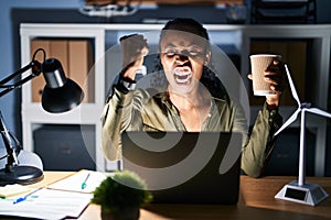 African woman working using computer laptop at night angry and mad raising fist frustrated and furious while shouting with anger