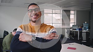 African young businessman standing in office with his arms crossed looking at camera with diverse colleagues talking in