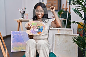 African young woman holding painter palette screaming proud, celebrating victory and success very excited with raised arms