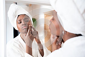 African young woman looking at pimple on face in mirror, worried unhappy check and touches her skin