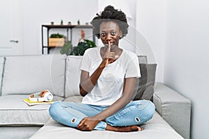 African young woman sitting on the sofa at home asking to be quiet with finger on lips