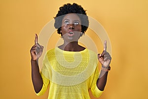 African young woman standing over yellow studio amazed and surprised looking up and pointing with fingers and raised arms