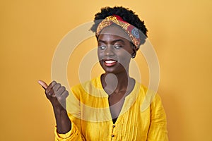 African young woman wearing african turban smiling with happy face looking and pointing to the side with thumb up