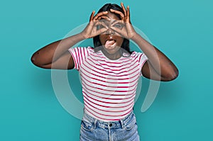 African young woman wearing casual striped t shirt doing ok gesture like binoculars sticking tongue out, eyes looking through