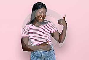 African young woman wearing casual striped t shirt smiling with happy face looking and pointing to the side with thumb up