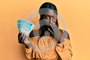 African young woman wearing doctor uniform holding 100 brazilian reals covering mouth with hand, shocked and afraid for mistake