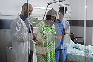 Afro american asisstant helping sick woman to stand up from bed holding intravenous IV fluid drip bag