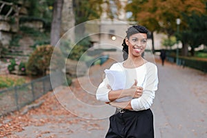 Afro american beautiful student walking with papers in autumn park, showing thumbs up.