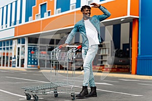Afro american black man with shopping cart in front of mall shopping mall, looks into the distance