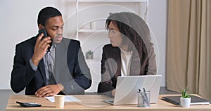 Afro american business man and woman colleague managers sitting at table working curly girl speaks secret into male ear