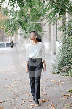 Afro american charming woman walking in town near green trees and building, wearing white blouse.