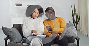 Afro american couple friends girl and guy sitting together in living room eating popcorn listening to music on radio