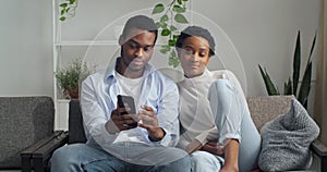 Afro american couple man and woman sitting together at home looking at phone screen using mobile device gadget to search