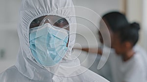 Afro american doctor scientist medical worker wears protective clothing hood transparent glasses mask looks at camera