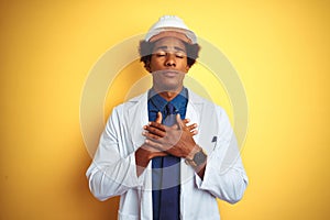 Afro american engineer man wearing white coat and helmet over isolated yellow background smiling with hands on chest with closed