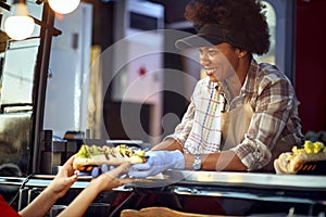 Afro-american female employee in fast food service giving sandwiches to a customer with a smile