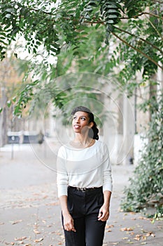 Afro american girl walking in town near green trees and building, wearing white blouse.