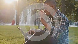 Afro american guy sits in park on grass makes online video call talking to laptop camera waving hello tells news