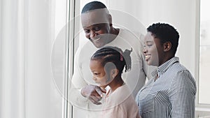 Afro american happy family, parents with little daughter looking out window standing at home expecting, black mixed race