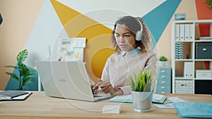 Afro-American lady in headphones listening to music and using laptop in modern office