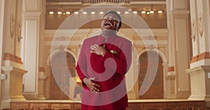 Afro american male performer in stylish red suit singing while standing at row of wooden pews in big hall. Talanted