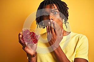 Afro american man with dreadlocks holding red currants over  yellow background cover mouth with hand shocked with shame