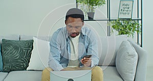 Afro american man holding plastic credit card in hand, entering payment data in computer application, sitting on sofa