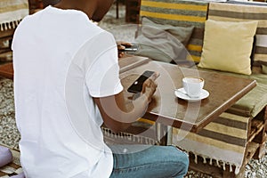 Afro american man sitting at table in cafe with cup of coffee on summer veranda, holding smartphone and entering number