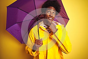 Afro american man wearing rain coat and umbrella standing over isolated yellow background serious face thinking about question,