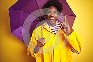 Afro american man wearing rain coat and umbrella standing over isolated yellow background surprised with an idea or question
