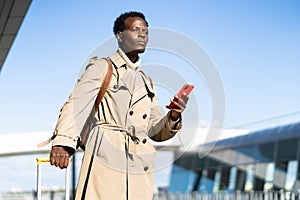 Afro-American millennial traveler man with yellow suitcase stands in airport terminal, holding phone, calling and looking for a