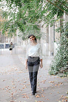 Afro american pretty woman walking in town near green trees and building, wearing white blouse.