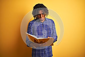 Afro american student man with dreadlocks reading book over isolated yellow background scared in shock with a surprise face,