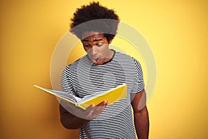 Afro american student man reading book standing over isolated yellow background scared in shock with a surprise face, afraid and