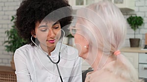 Afro american woman doctor listens to breath of senior woman using stethoscope while sitting on sofa at home